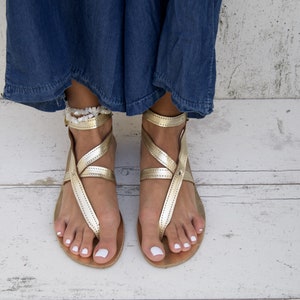 May include: Gold leather sandals with ankle straps and a white beaded ankle bracelet. The sandals are on a white wooden surface.