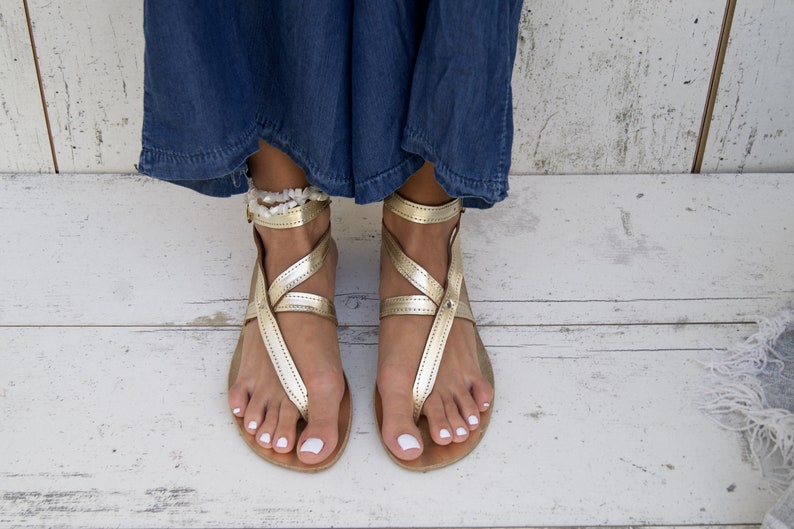 May include: Gold leather sandals with ankle straps and a white beaded ankle bracelet. The sandals are on a white wooden surface.