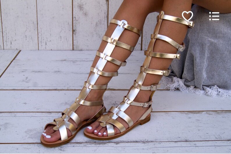 May include: Gold gladiator sandals with multiple straps and buckles. The sandals are knee-high and have a flat sole.