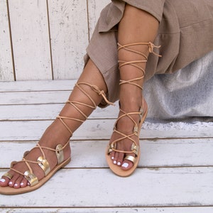 May include: A pair of gold leather sandals with tan laces that tie up the legs. The sandals have a flat sole and are worn by a person sitting on a white wooden surface.