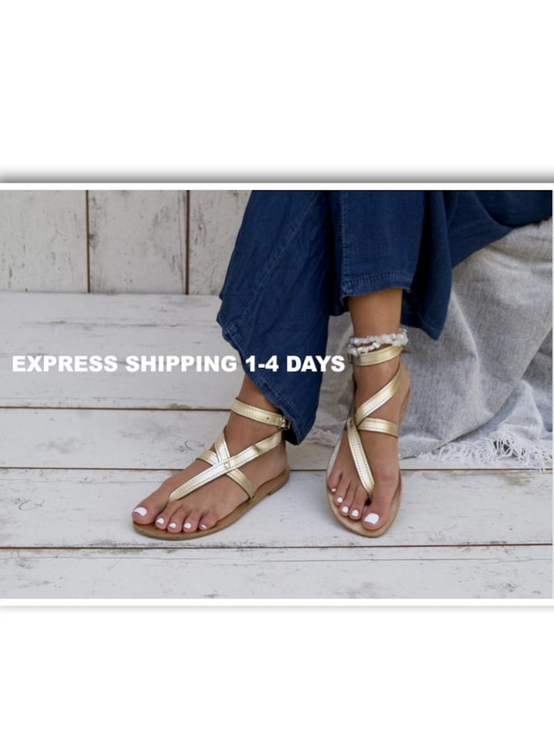 May include: Gold leather sandals with ankle straps and a criss-cross design. The sandals are worn on a wooden surface.