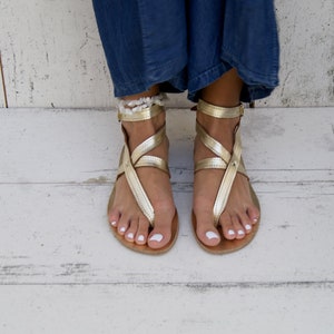 May include: Gold leather sandals with ankle straps and a criss-cross design. The sandals have a toe loop and are worn on a pair of feet.