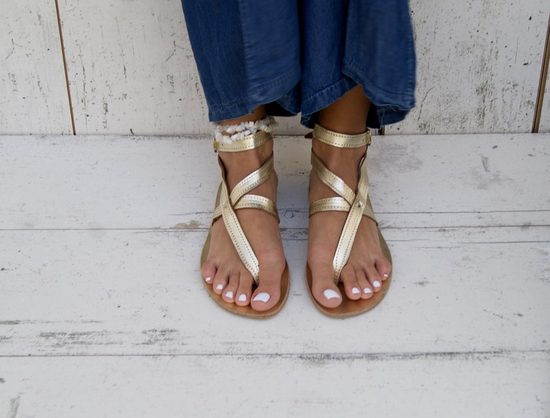 May include: Gold leather sandals with ankle straps and a criss-cross design. The sandals have a toe loop and are worn on a pair of feet.