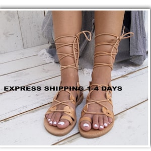 May include: Tan leather sandals with a strappy design that wraps around the legs. The sandals have a flat sole and are shown on a light wood surface. The text "EXPRESS SHIPPING 1-4 DAYS" is visible.