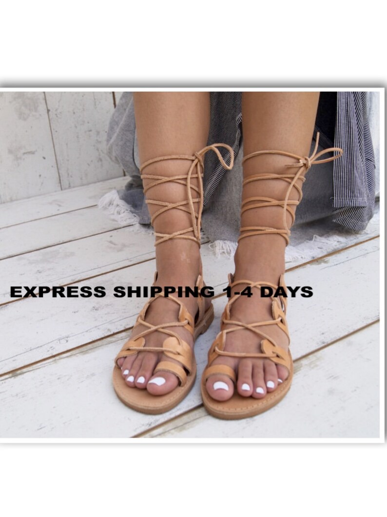May include: Tan leather sandals with a strappy design that wraps around the legs. The sandals have a flat sole and are shown on a light wood surface. The text "EXPRESS SHIPPING 1-4 DAYS" is visible.