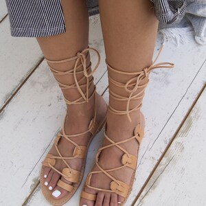 May include: Tan leather sandals with multiple straps that wrap around the ankles and lower legs. The sandals have a flat sole and are displayed on a white wooden surface. The sandals are a neutral color and appear to be handmade.