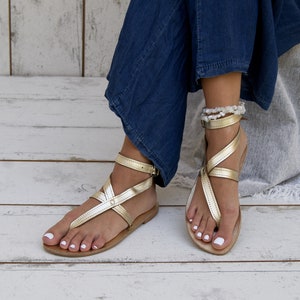 May include: A pair of gold leather sandals with ankle straps. The sandals have a simple design with a criss-cross strap pattern. The sandals are worn by a person with white nail polish.