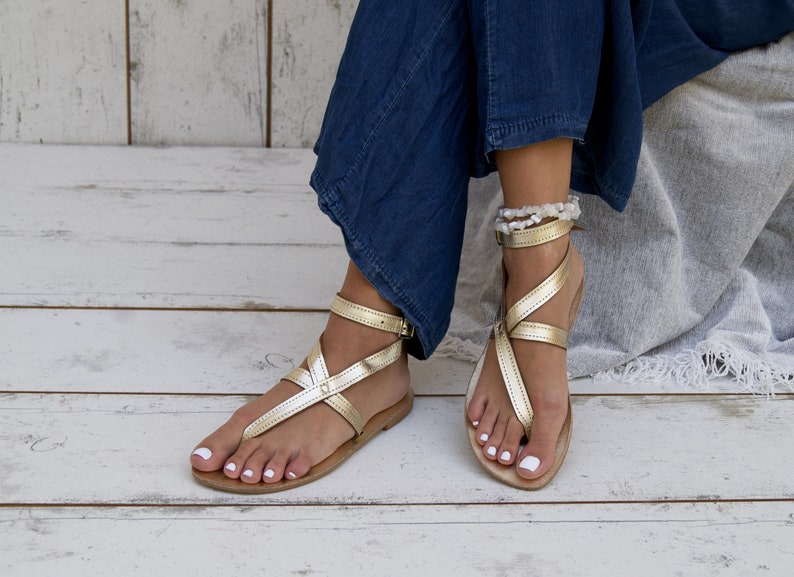 May include: A pair of gold leather sandals with ankle straps. The sandals have a simple design with a criss-cross strap pattern. The sandals are worn by a person with white nail polish.