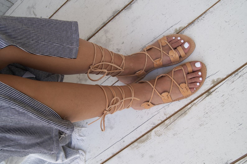 May include: Tan leather gladiator sandals with lace-up design. The sandals feature a toe loop and ankle straps. The image shows the sandals on a white wooden surface. The sandals are a neutral color.