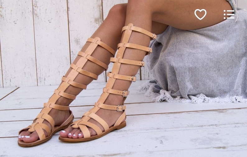 May include: A pair of tan leather gladiator sandals with multiple straps and buckles. The sandals are worn by a person with bare legs and are on a light wood floor.