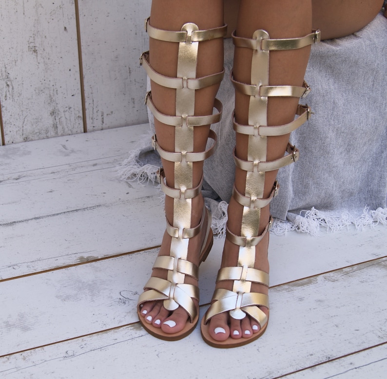 May include: Gold gladiator sandals with multiple straps and buckles. The sandals are made of leather and have a flat sole.
