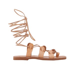 May include: A single, light brown leather sandal with a flat sole. The sandal features multiple straps across the foot and a long, wraparound ankle tie. The design includes a toe loop and a series of vertical straps.