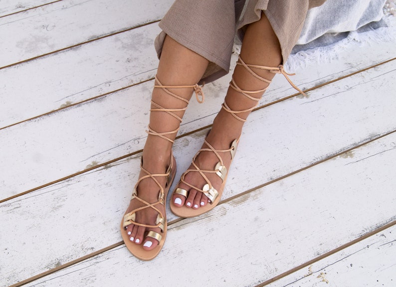 May include: A pair of tan leather sandals with gold accents. The sandals have laces that tie up the leg. The sandals are on a white wooden surface.