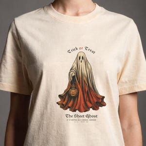 Peut inclure: Un t-shirt couleur crème avec un motif représentant un fantôme drapé dans un linceul, tenant une citrouille d'Halloween. Le texte indique "Trick or Treat", "The Sheet Ghost" et "IT STARTED AS A BURIAL SHROUD". Le drap du fantôme passe du blanc à l'orange.