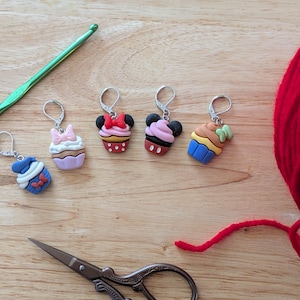 Peut inclure: Cinq marqueurs de points de couture en forme de cupcake colorés avec des fermoirs argentés. Les marqueurs représentent Mickey Mouse, Minnie Mouse, un cupcake bleu, un cupcake rose et un cupcake vert. Les marqueurs sont présentés sur une surface en bois avec un peloton de laine rouge et un crochet vert.