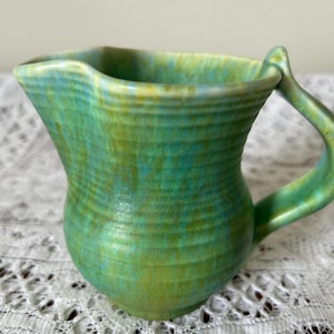Könnte beinhalten: Handgefertigter Keramikkrug mit einer einzigartigen, organischen Form. Der Krug ist in Grün- und Blautönen glasiert, wodurch ein strukturiertes, gesprenkeltes Aussehen entsteht. Der Griff krümmt sich anmutig vom Körper. Dieser dekorative Krug ist ideal zum Servieren von Getränken.