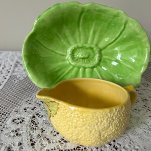 Op de afbeelding: Een heldergroene, bloemvormige keramische schaal en een kleine gele creamer met een gestructureerd oppervlak. De creamer heeft een handvat en een tuit. De schaal staat op de achtergrond en de creamer op de voorgrond. De items staan op een wit kanten tafelkleed.