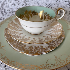 Può includere: Un servizio da tè vintage con una tazza bianca con interno verde menta e design a foglie d'oro, appoggiata su un piattino abbinato e un piatto più grande. Il set ha finiture dorate e motivi floreali.