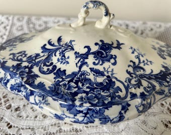 Puede incluir: Un plato de servir de porcelana blanca con un patrón floral azul y blanco. El plato tiene una tapa con un asa.
