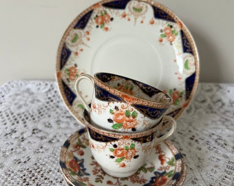 Könnte beinhalten: Zwei antike Teetassen und eine Untertasse mit einem floralen Muster in Orange-, Grün- und Blautönen. Die Tassen haben einen goldenen Rand und die Untertasse einen blauen und goldenen Rand.