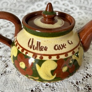 May include: A small, brown ceramic teapot with a green and yellow floral design. The teapot has a lid with a knob on top and the words "Wulcea ave a" written on the side.