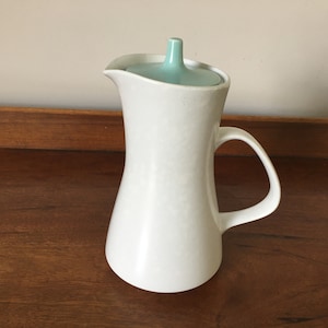 May include: White ceramic pitcher with a light blue lid. The pitcher has a wide base and a narrow neck. It has a handle on the side.