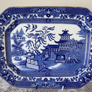 Puede incluir: Una fuente de cerámica octogonal azul y blanca con borde dorado. La fuente presenta una escena detallada del patrón Willow con pagodas, un puente y pájaros. El borde tiene un diseño floral y geométrico.