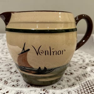 May include: A brown and cream ceramic jug with a green stripe and a brown handle. The jug has a painted image of a sailboat and the word "Ventnor" on the side.