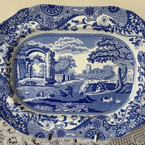May include: A blue and white ceramic platter with an oval shape and a detailed landscape design. The border has floral and scroll patterns. The central image shows a rural scene with buildings, trees, and figures, a classic decorative item.