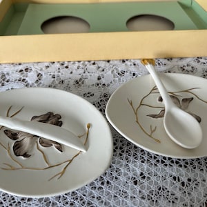 May include: Two white ceramic dishes with gold accents and a floral design. Each dish has a matching spoon, one with a gold handle and the other with a white handle.