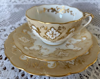 May include: A three-piece tea set with a delicate floral design in gold and white on a cream background. The set includes a cup, saucer, and a larger plate.