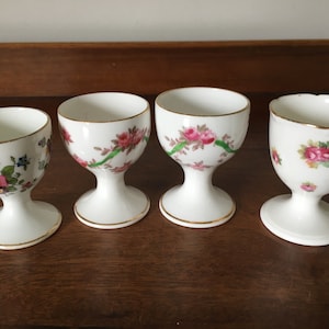 May include: Four white ceramic egg cups with floral designs. The egg cups have a gold trim and are decorated with pink and green roses.