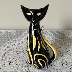 May include: A black ceramic cat figurine with yellow and white accents. The cat has a sleek, elongated body, pointed ears, and a curved tail. The yellow design creates a flame-like pattern on the cat's body. The eyes are yellow.