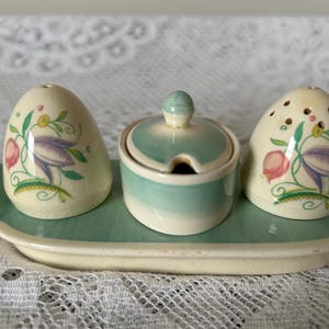 May include: A vintage ceramic salt and pepper shaker set with a matching lidded sugar bowl, all resting on a rectangular tray. The set features a floral design with purple, pink, and green accents. The tray and bowls are light green.