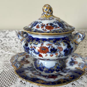 May include: A vintage porcelain soup tureen with a lid, handles, and a matching saucer. The tureen and saucer feature a detailed floral pattern in shades of blue, orange, and gold. The rim and handles are accented with gold trim.