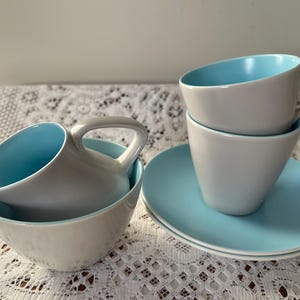 May include: A vintage tea set with a light grey and blue colour scheme. The cups and creamer have a light blue interior and a light grey exterior. The cups are stacked on a saucer with a light blue rim.