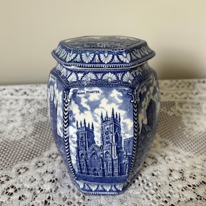 Puede incluir: Un tarro de cerámica hexagonal con tapa, que presenta un patrón de transferencia en azul y blanco. El diseño representa la catedral de York Minster con el texto "YORK MINSTER" impreso sobre ella. El tarro tiene bordes decorativos.