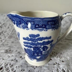 Puede incluir: Una cremera de cerámica blanca y azul con asa y boquilla. La cremera presenta un diseño de patrón Willow con una pagoda, árboles y un puente. La cremera está sobre un mantel de encaje blanco.