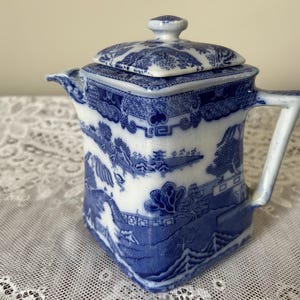 May include: A square-shaped ceramic teapot featuring a blue and white willow pattern. The teapot has a matching lid with a small knob handle, a short spout, and a single angular handle. The surface is decorated with detailed landscape scenes.