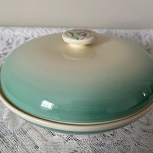 Könnte beinhalten: Ein zweiteiliges Keramik-Serviergeschirr mit Deckel. Das Geschirr wechselt von einer cremefarbenen Farbe oben zu einem türkisgrün am Boden. Der Deckel hat einen kleinen, runden Griff mit floralem Design.