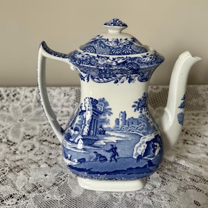 Peut inclure: Une théière en céramique blanche ornée d'un motif bleu de type transfert représentant un paysage avec des ruines, un château et des personnages. La théière comprend un couvercle assorti à bordure florale, une anse courbée et un bec verseur.