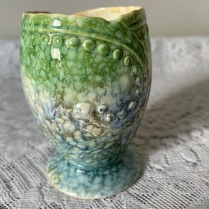 Peut inclure: Un coquetier en céramique avec un glaçage texturé et moucheté dans les tons de vert, de bleu et de crème. Le coquetier a un corps arrondi et une petite base en piédestal. Des points en relief ornent le côté.