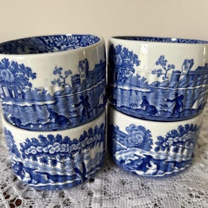 Puede incluir: Cuatro cuencos de cerámica blancos y azules con un diseño de paisaje. Los cuencos presentan un patrón de transferencia azul y blanco que representa una escena fluvial con figuras, árboles y edificios. Los cuencos están apilados sobre un mantel de encaje blanco.