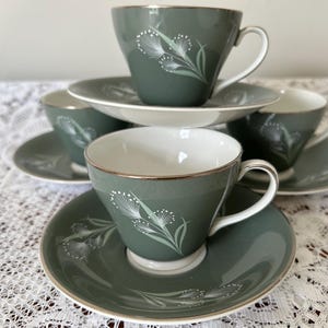 Puede incluir: Un juego de seis tazas y platillos vintage. Las tazas y los platillos son de color verde apagado con un diseño floral blanco y ribete dorado. Las tazas están apiladas, con los platillos debajo.