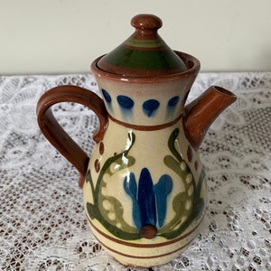 Op de afbeelding: Een klein, bruin aardewerken theepotje met een deksel. De theepot is versierd met groene en blauwe bloemenmotieven.