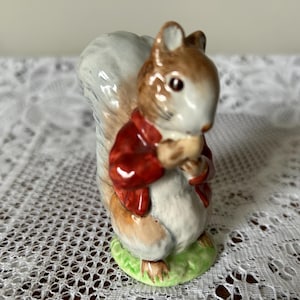 May include: A small porcelain figurine of a squirrel wearing a red jacket and holding a nut. The squirrel has a bushy tail and is standing on a green base.
