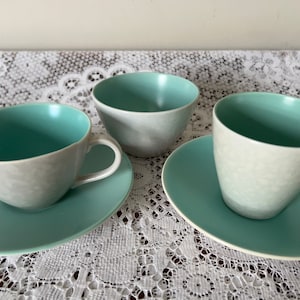 May include: Three ceramic teacups and saucers in a light grey and teal colour scheme. The cups and saucers have a textured surface.
