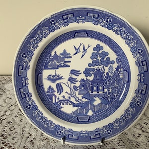 May include: A white ceramic dinner plate with a blue and white willow pattern. The plate features a traditional Chinese landscape scene with a pagoda, a bridge, and a willow tree.