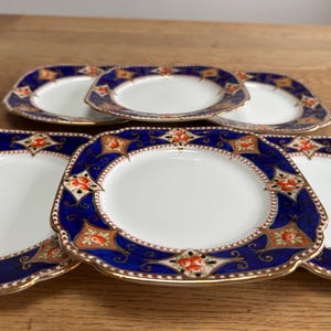 Puede incluir: Juego de seis platos cuadrados de cerámica con borde azul y dorado y centro blanco. Los platos tienen un diseño floral en naranja y rojo.