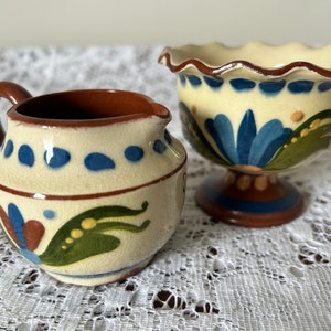 Puede incluir: Un juego de dos cuencos pequeños de cerámica pintados a mano con una base crema y diseños florales azules, verdes y marrones. Los cuencos están sobre un mantel de encaje blanco.